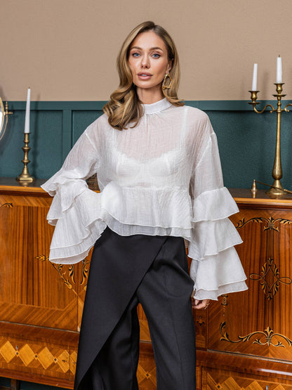 Woman wearing a white blouse with ruffled sleeves and black pants in an elegant room.
