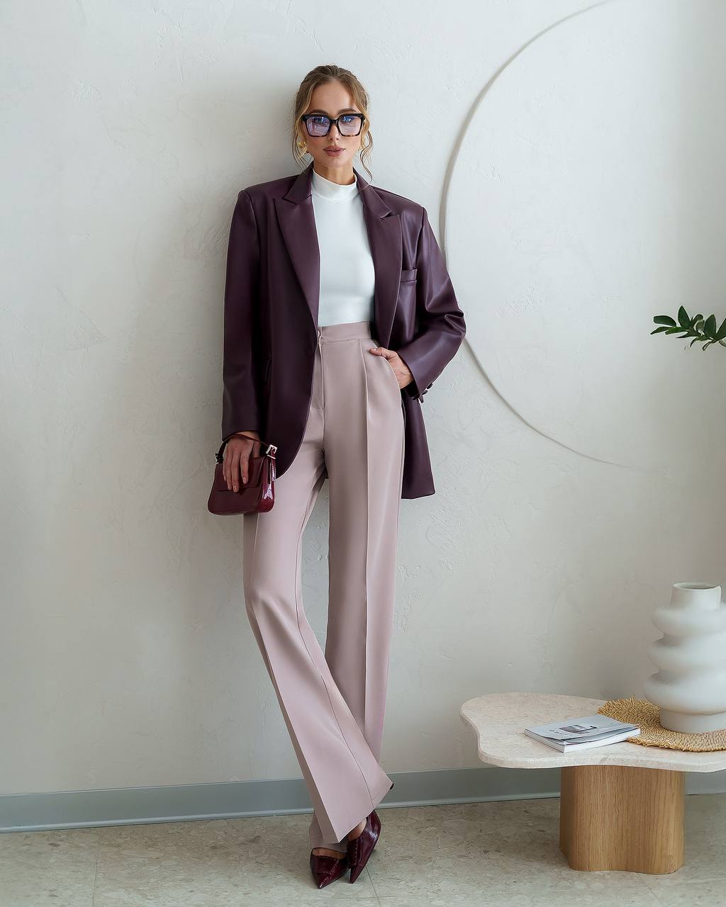 Woman in a stylish outfit with a purple blazer and pink pants standing in a modern room.
