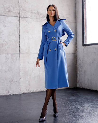 Woman wearing a blue trench coat with gold buttons against a gray concrete wall.