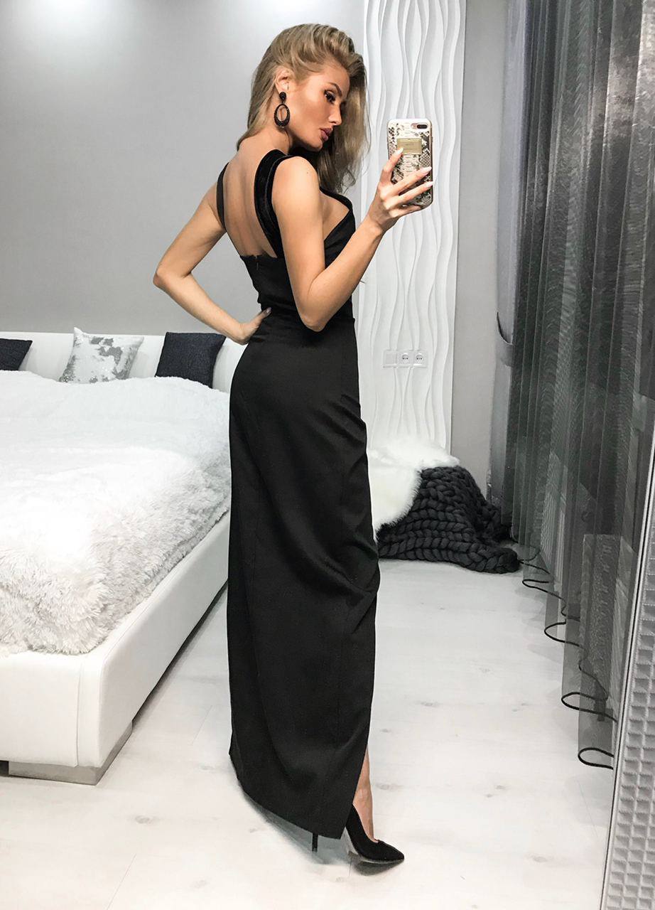 A woman takes a mirror selfie in a bedroom wearing a floor-length black evening gown with a low open back and a high slit, paired with black pointed-toe heels.