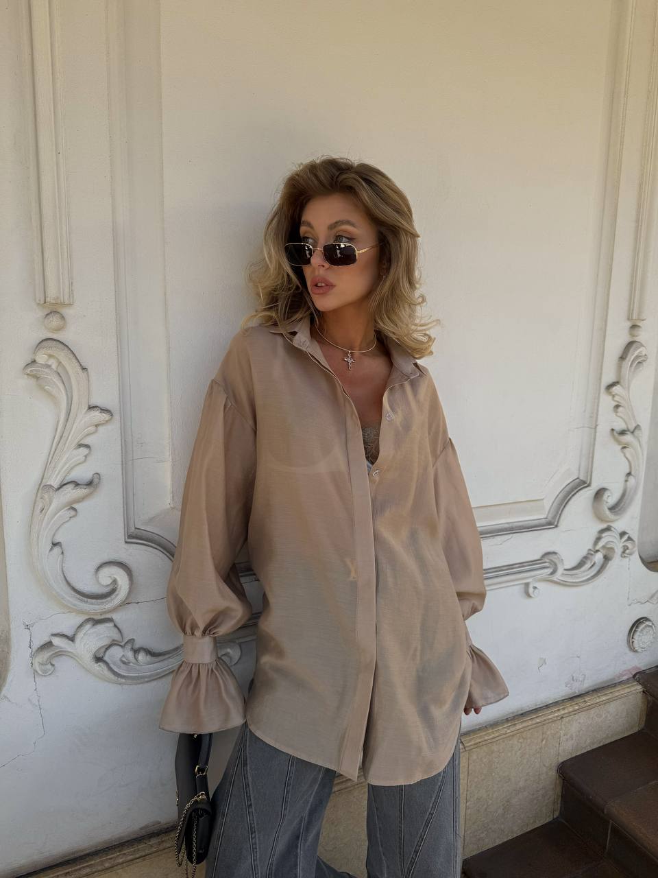 Woman wearing a beige shirt and sunglasses against an ornate wall.