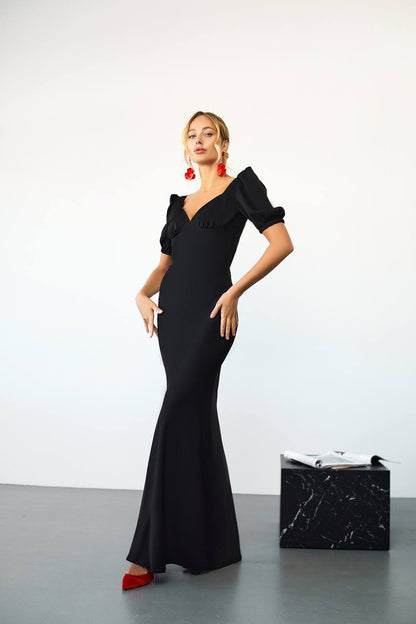 Woman in a black evening gown with red shoes and earrings standing next to a black handbag on a light gray floor.