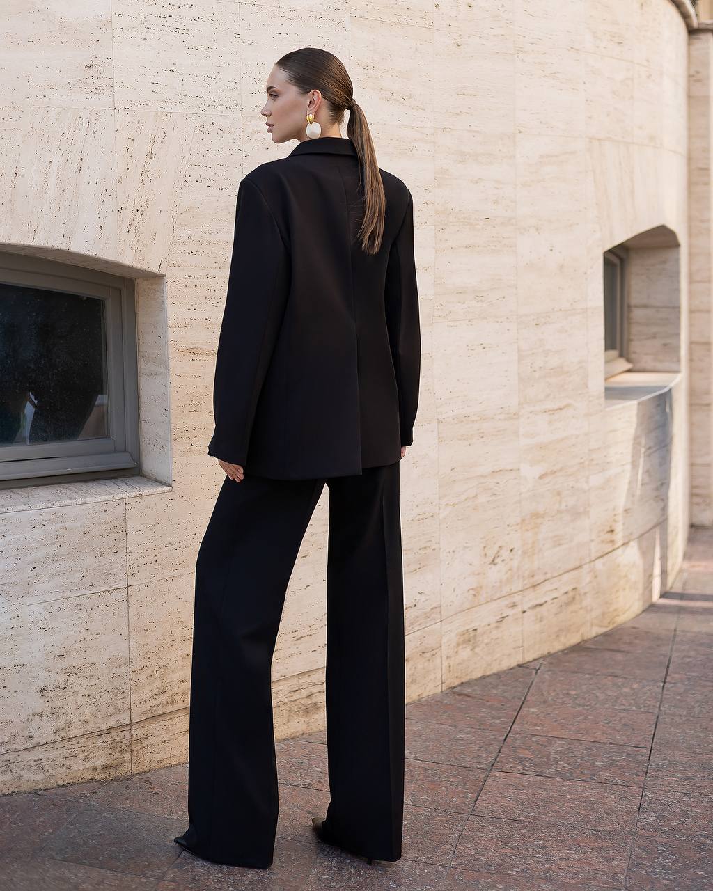 Woman in a black suit standing against a beige stone building.