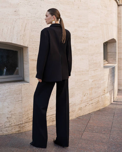 Woman in a black suit standing against a beige stone building.