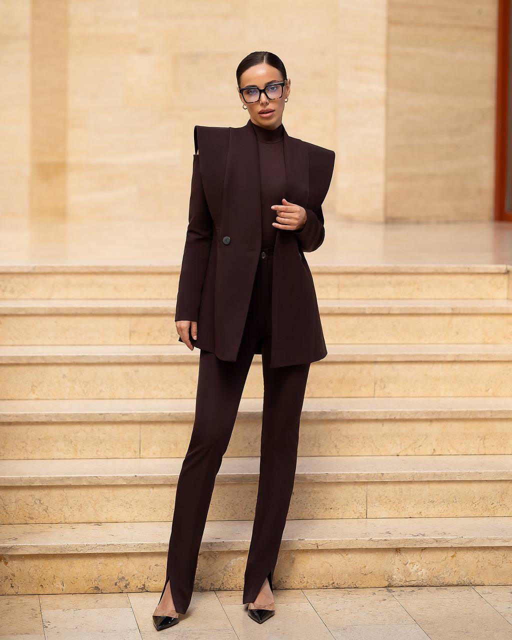 A model stands on beige stone steps wearing a dark brown structured blazer with dramatic shoulder pads, matching slim-fit trousers, a high-neck top, and black pointed heels.
