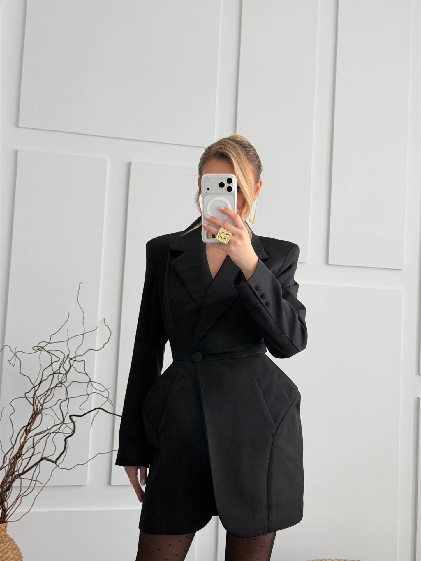 A woman takes a mirror selfie wearing a black structured blazer-style mini dress with a wrap waist and oversized sleeves, standing against a white paneled wall.