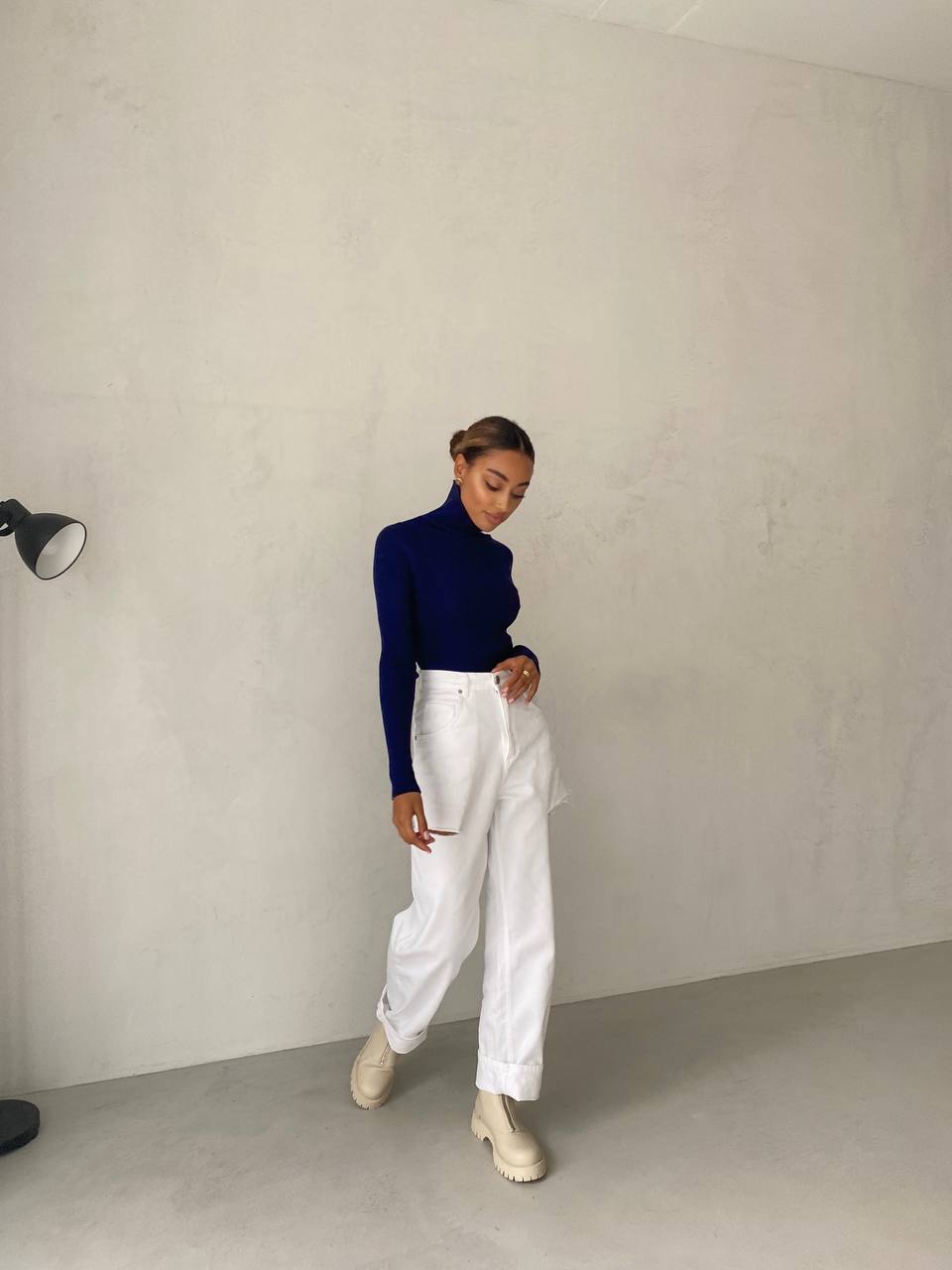 Person wearing a navy sweater and white pants against a plain wall.