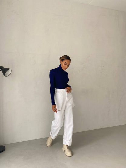 Person wearing a navy sweater and white pants against a plain wall.
