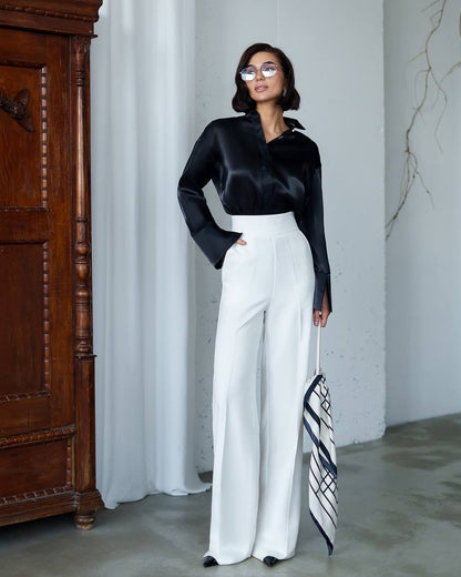 Woman in black satin blouse and white pants standing in a room with wooden furniture and white curtains.