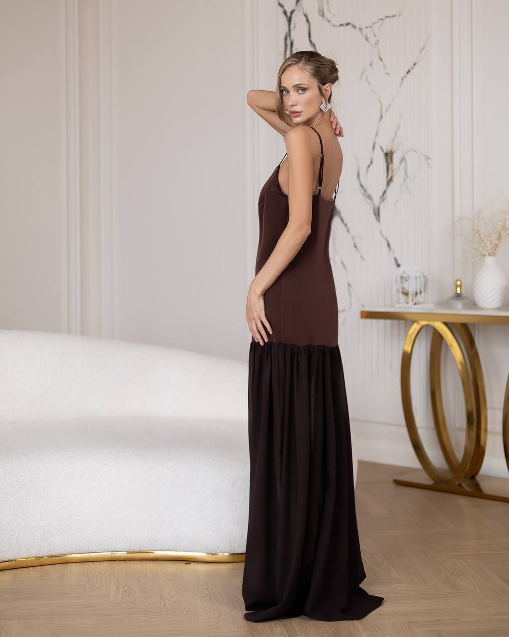 Woman in a long brown dress standing in a stylish room with marble walls and modern decor.