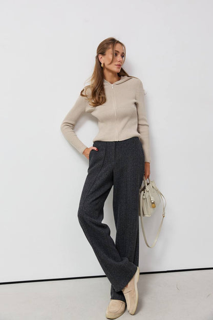 Woman wearing a beige sweater and gray pants against a white background