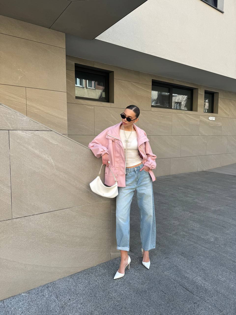 Woman in a pink jacket and light blue jeans standing against a modern building.