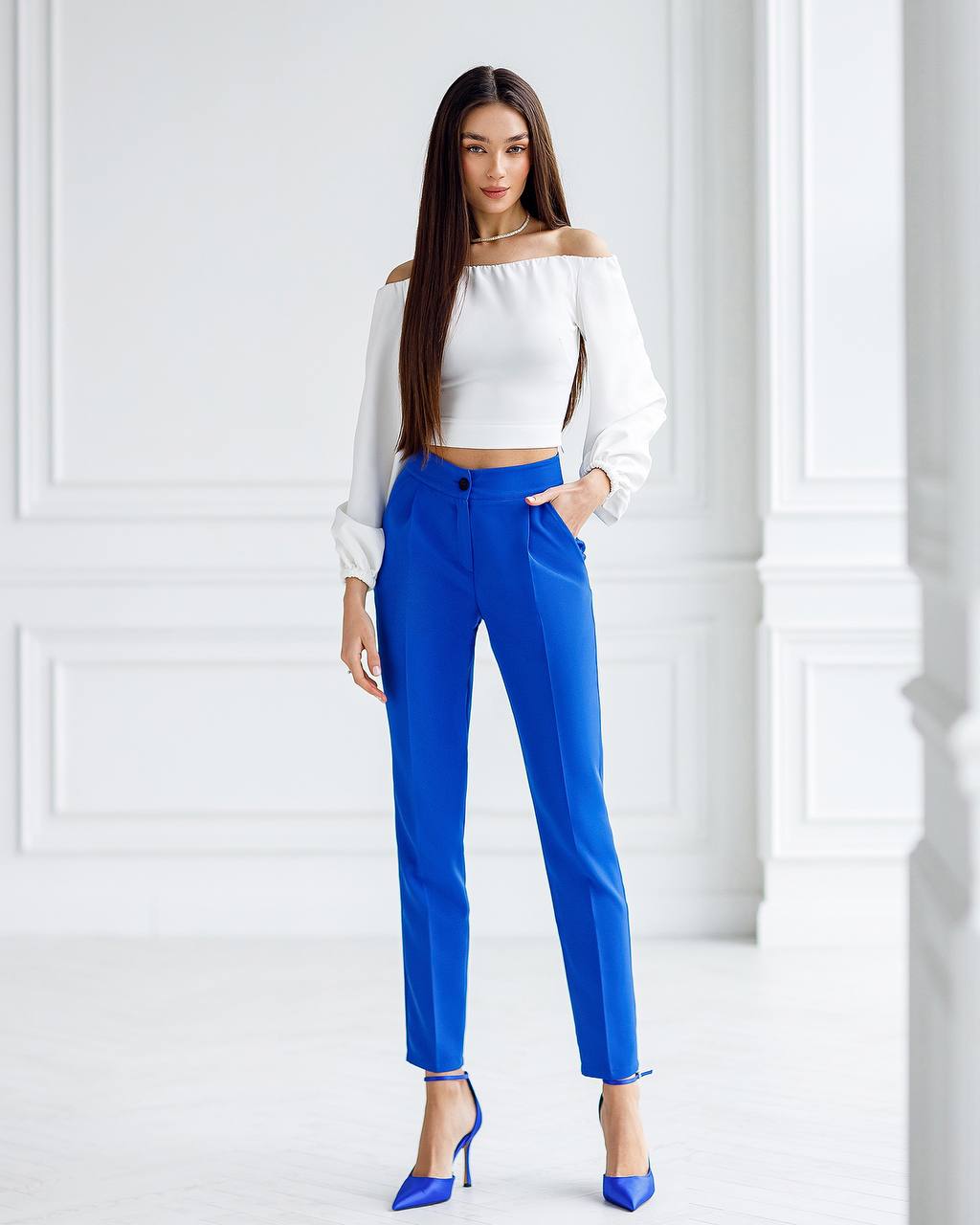 Woman wearing a white off-shoulder top and blue pants against a white wall.
