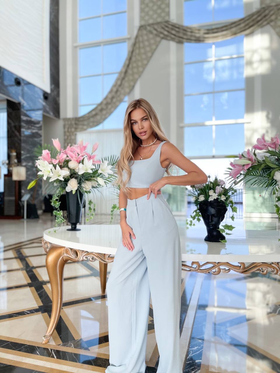 Woman in a light blue top and white pants standing in a luxurious room with large windows and decorative table.