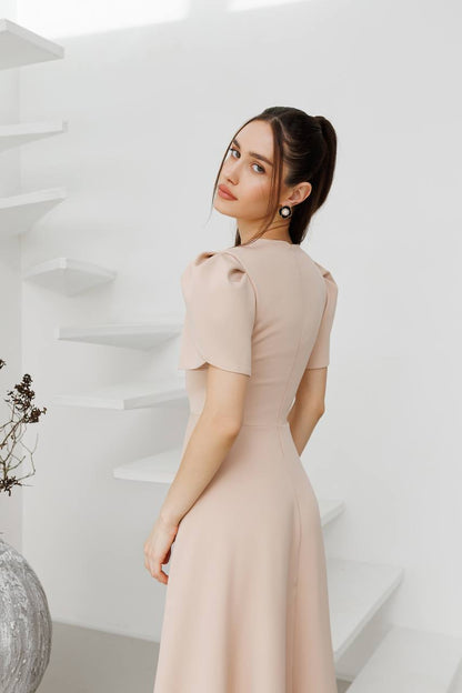 Woman in a beige dress standing in a minimalistic room with white walls and a vase.