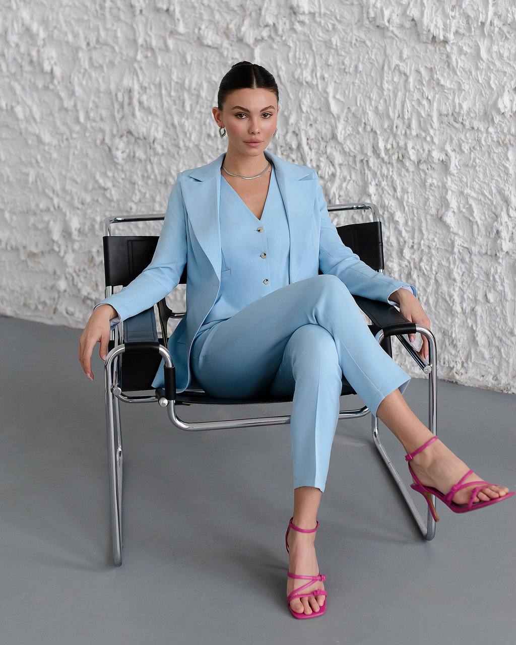 Woman in a light blue suit sitting on a chair against a textured white wall.