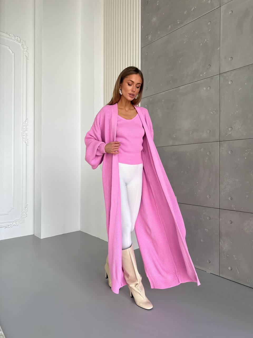 Woman wearing a long pink coat over a pink top and white pants in a minimalistic setting.