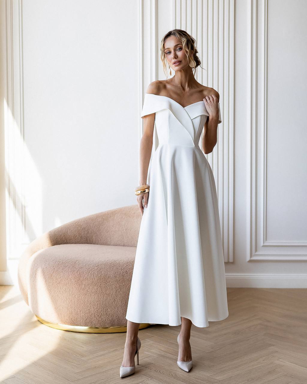 Woman in a white off-shoulder dress standing in a stylish room with a large window.