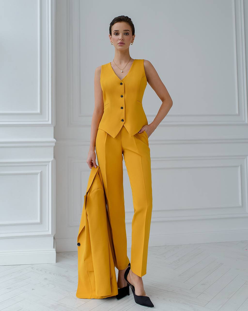 Woman wearing a yellow sleeveless top and wide-leg pants against a white paneled wall.