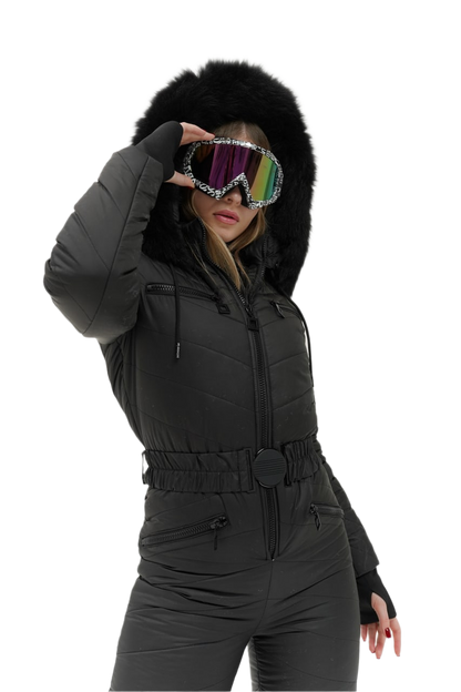 Person wearing a black snowsuit with a fur-lined hood, holding colorful goggles.