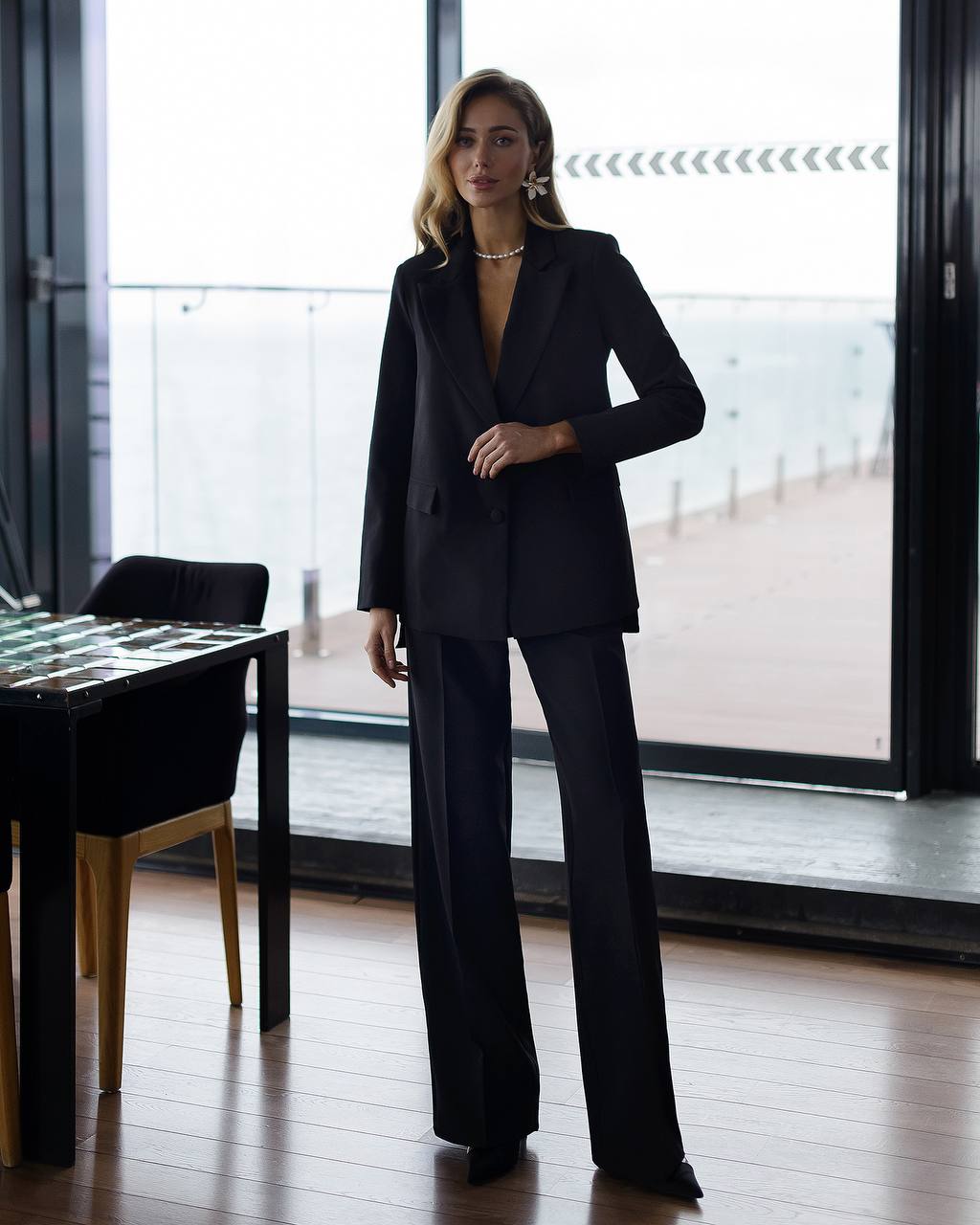 Woman in a black suit standing in a modern interior setting with large windows.