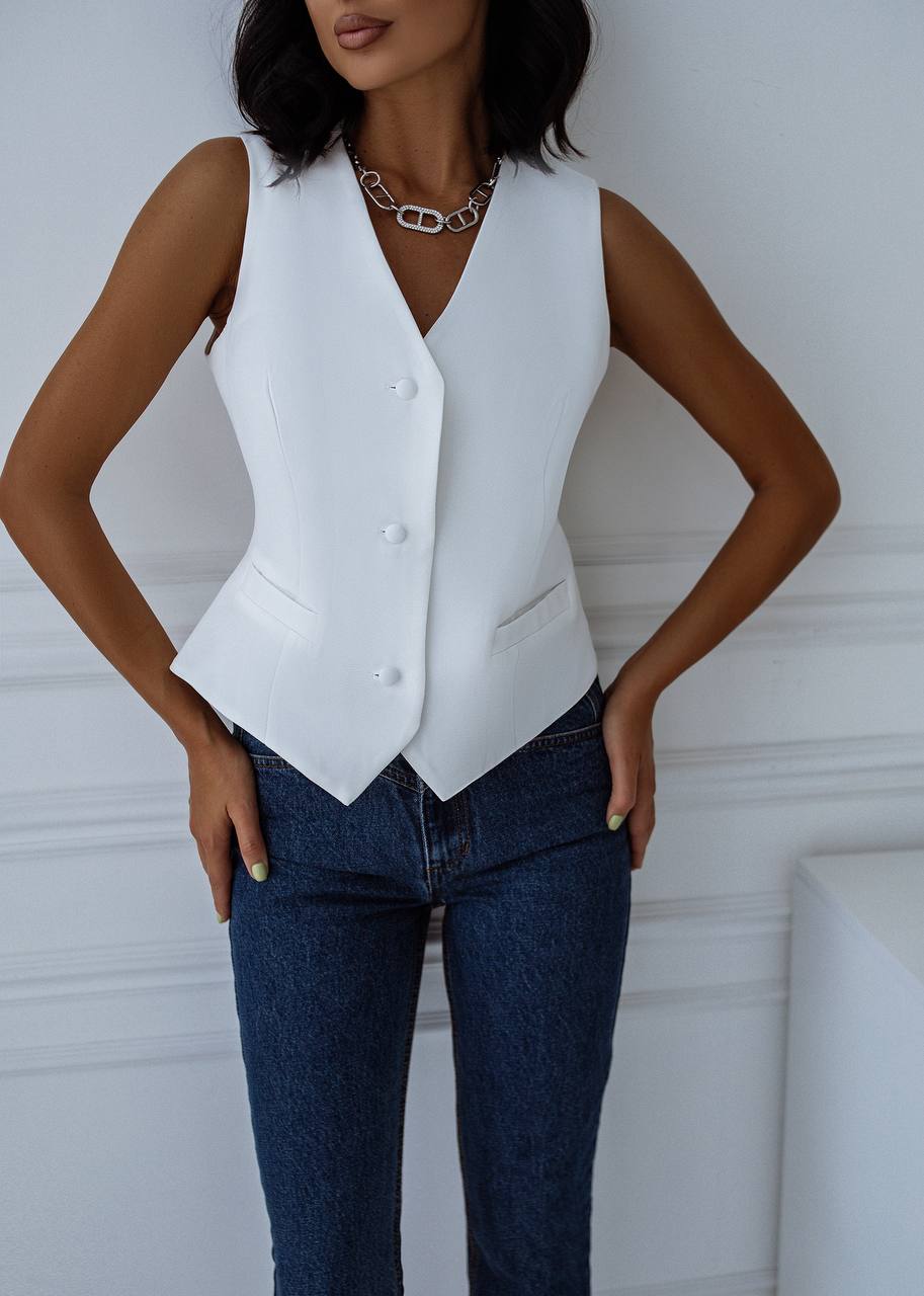 Person wearing a white vest over a black top with blue jeans against a white wall.