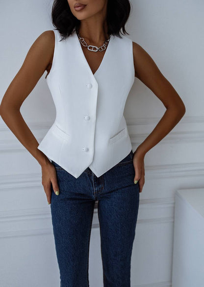 Person wearing a white vest over a black top with blue jeans against a white wall.