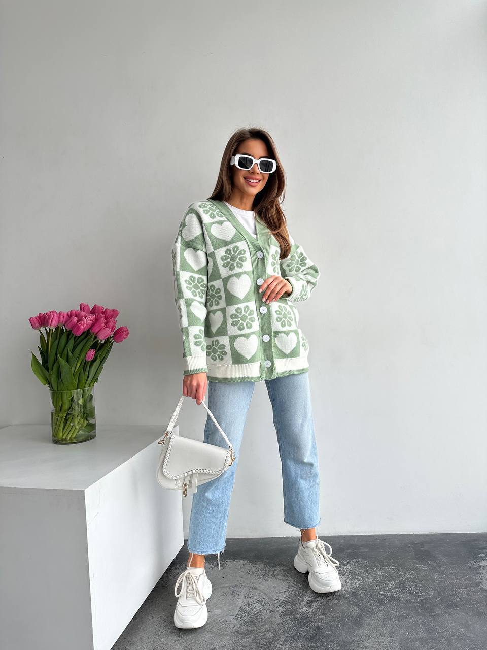 Woman wearing a green patterned sweater with a vase of pink tulips on a white surface.