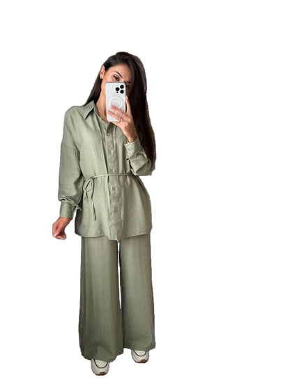 Person wearing a green outfit with wide-leg pants, holding a phone, and standing next to a small handbag on a white background.