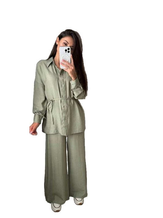 Person wearing a green outfit with wide-leg pants, holding a phone, and standing next to a small handbag on a white background.