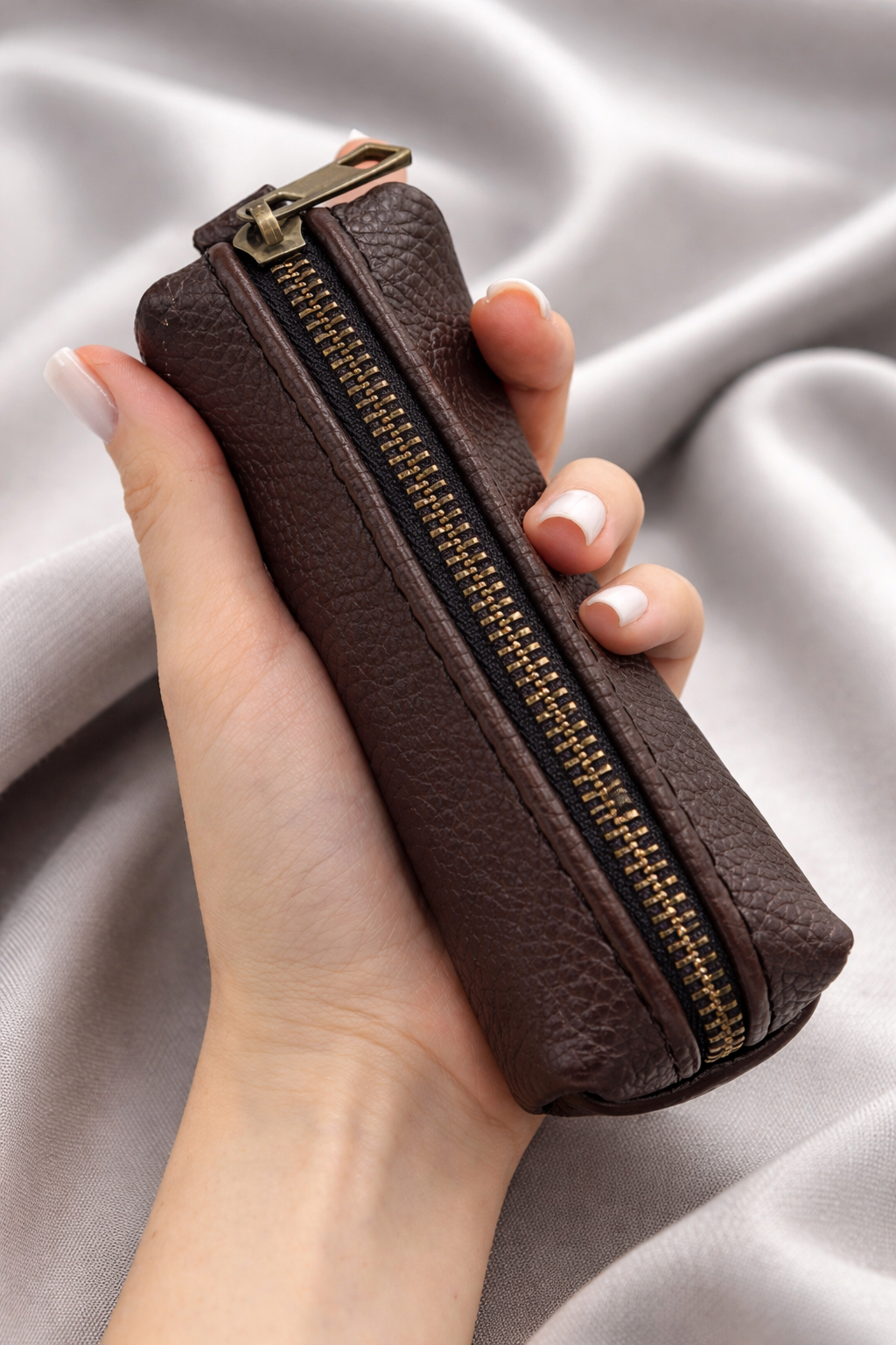A hand holding a small brown faux-leather zip pouch with a gold-tone zipper, set against a draped light gray fabric background.