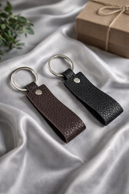 Two pebbled leather keychain loops, one dark brown and one black, with silver metal key rings, displayed on draped silver satin fabric with a brown gift box in the background.