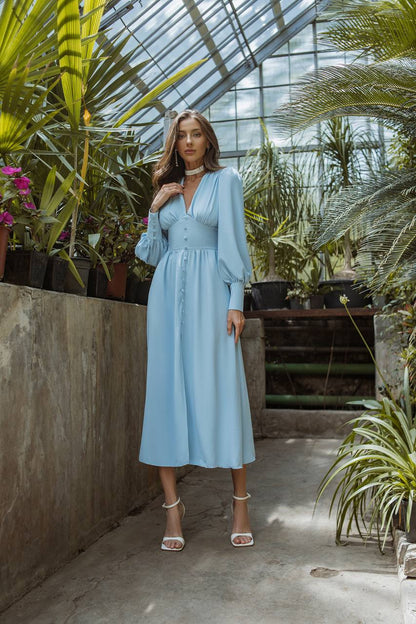 Woman in a light blue dress standing in a greenhouse filled with plants.