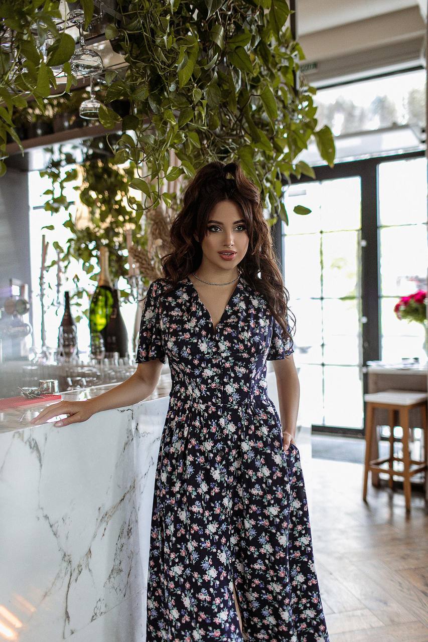 Woman in a floral dress standing in a modern interior setting with plants and decor.