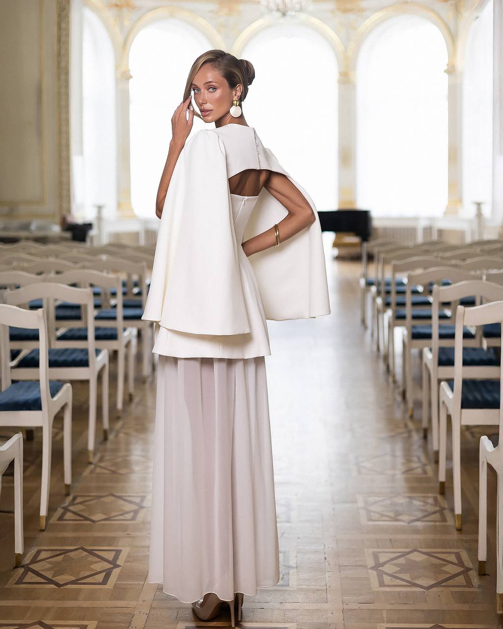 Woman in a white outfit standing in an elegant hall with arched windows.