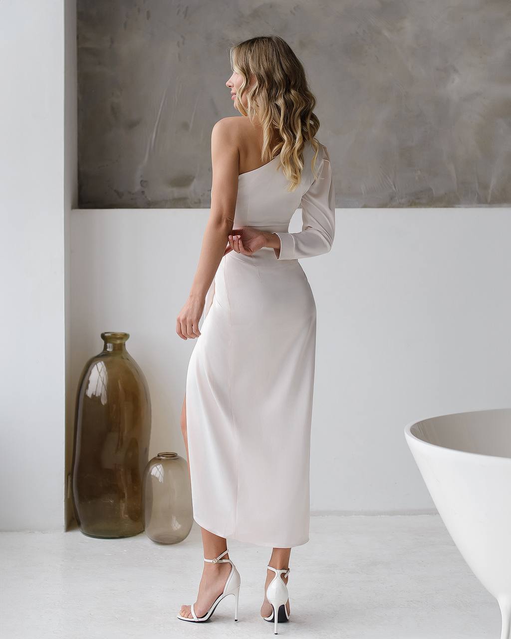 Woman in a white one-shoulder dress standing in a minimalistic room with decorative vases.
