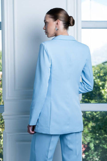 Person wearing a light blue suit standing in front of a window with greenery outside.