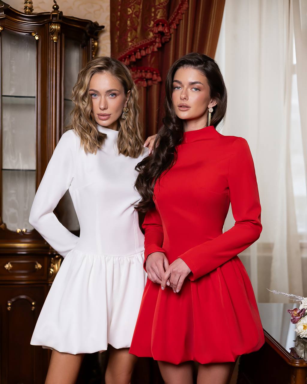 Two women modeling long-sleeve mini dresses in an indoor setting. The woman on the left wears a white dress, and the woman on the right wears a bright red dress. A dark wood cabinet and cream curtains are visible in the background.