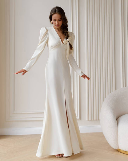 Woman in a white wedding dress standing in a room with light-colored walls and a chair.