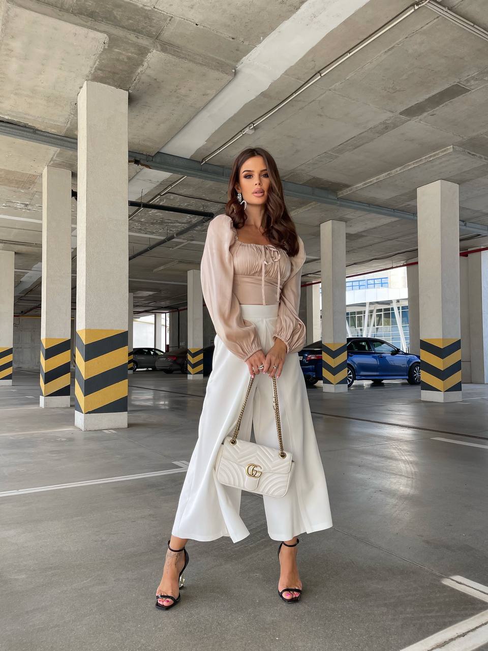 Woman in a beige top and white pants holding a white handbag in a parking garage.