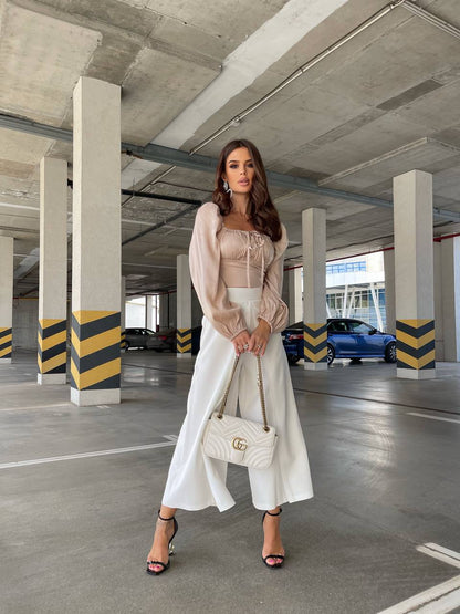Woman in a beige top and white pants holding a white handbag in a parking garage.