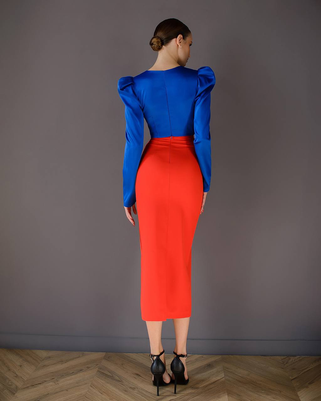 Woman wearing a blue top and red skirt against a gray wall.