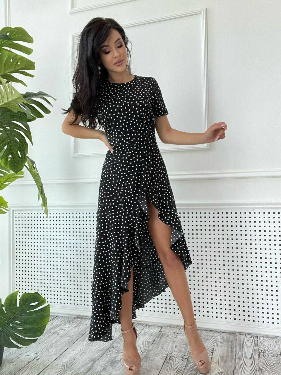 Woman wearing a black polka dot dress with a high-low hem in a room with white walls and a plant.