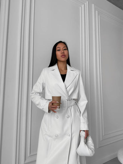 Woman in a white trench coat holding a coffee cup against a gray wall.
