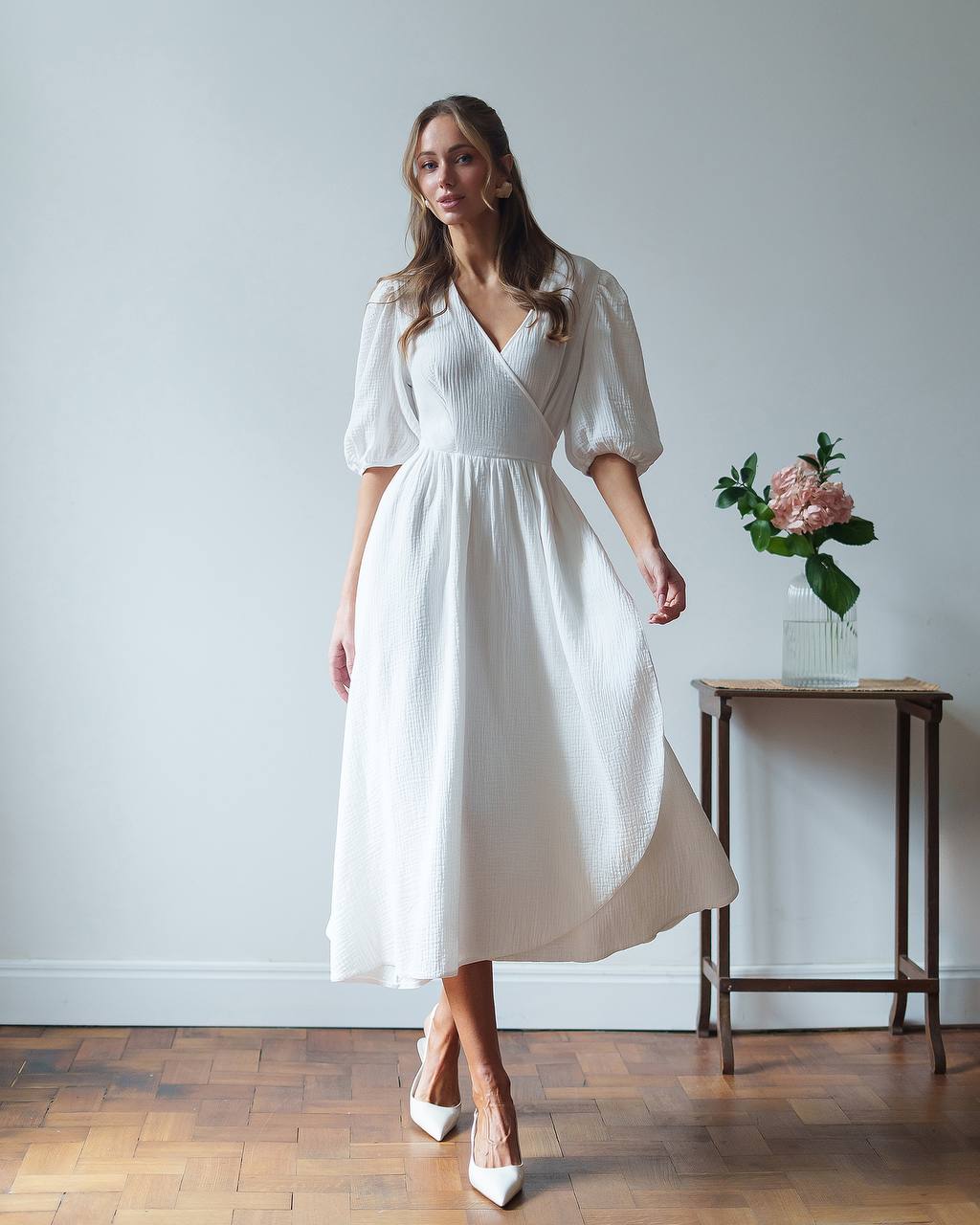 Woman wearing a white dress standing in a room with a wooden floor and a small table with flowers.