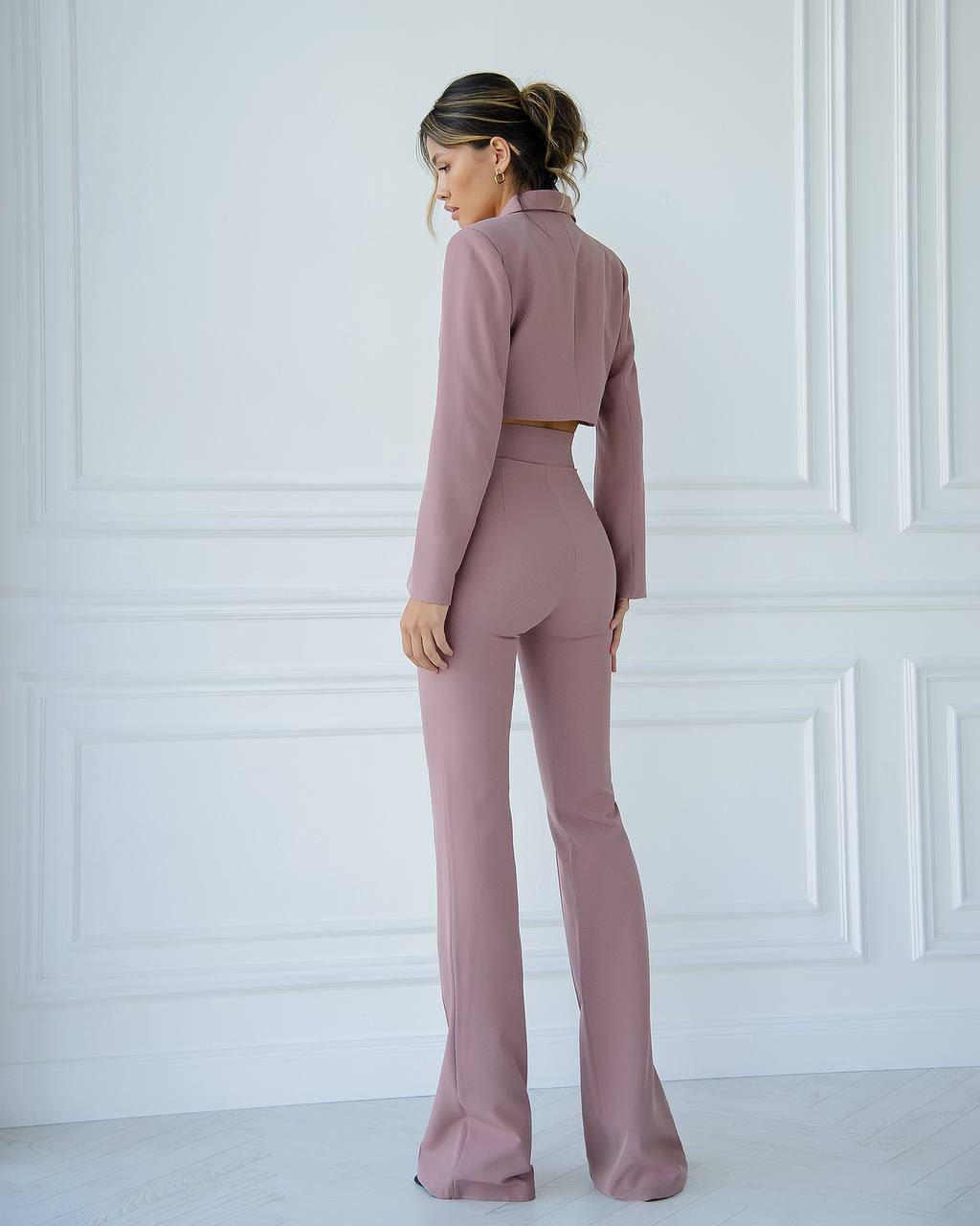 Woman wearing a pink suit standing against a white paneled wall.