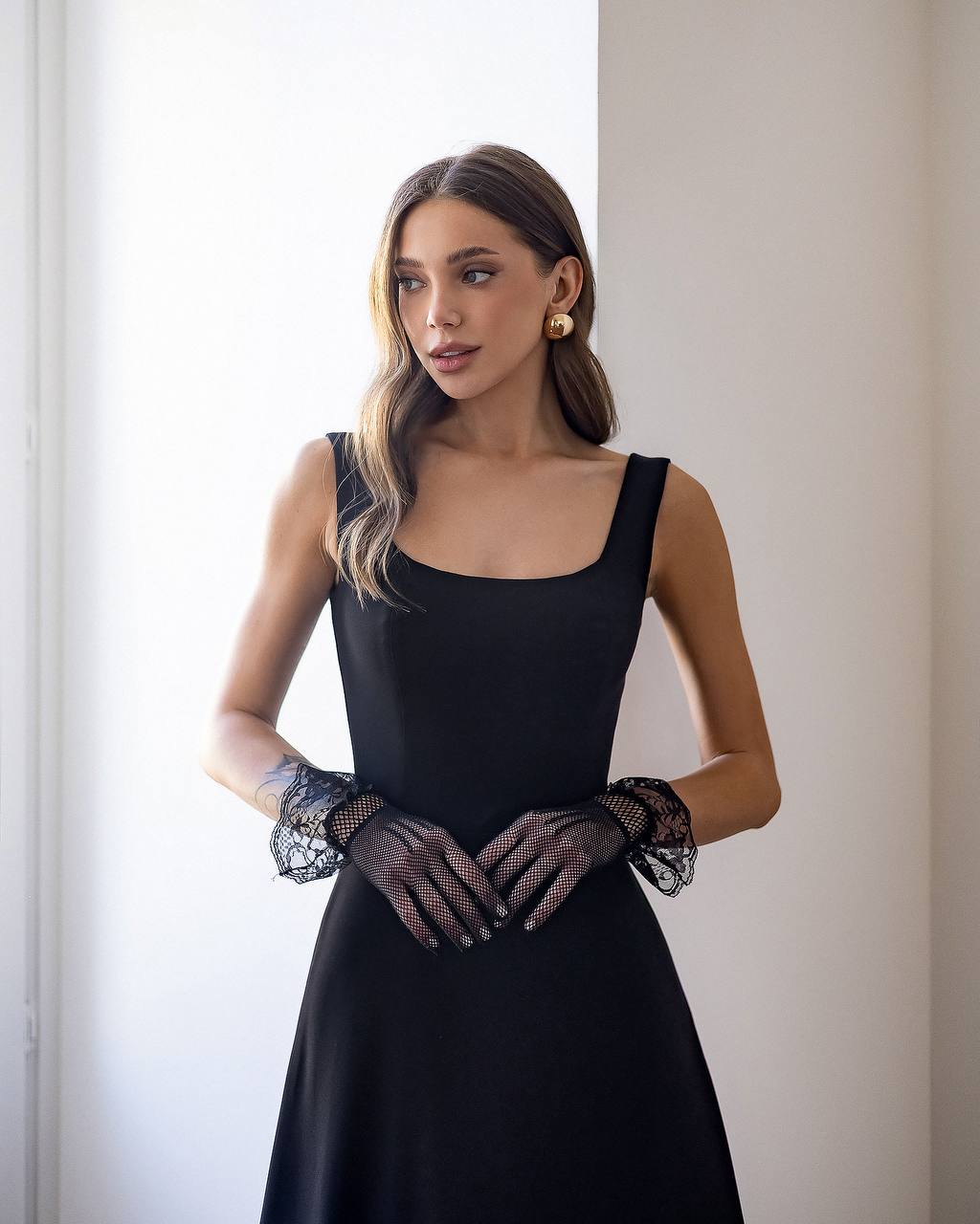 Woman wearing a black dress with sheer sleeves and gloves against a neutral background