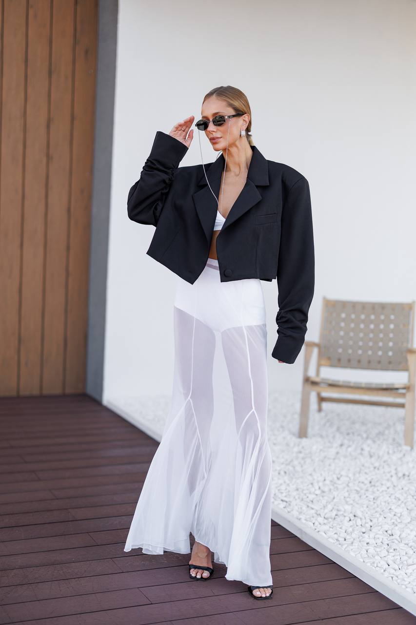 A woman models a black cropped blazer over a white top and a sheer white maxi skirt, standing on a dark wooden deck next to a white wall with a woven chair.