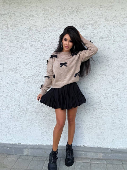 Woman wearing a beige sweater with black bow details and a black pleated skirt against a white wall.