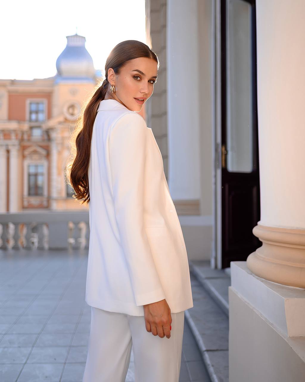 Woman in a white outfit standing in an urban setting with classical architecture.